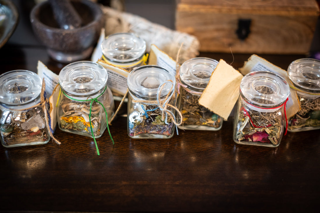 Row of glass spell jars containing herbs and crystals on a rustic wooden table, with a vintage wooden box and a mortar and pestle set in the background, symbolizing traditional witchcraft and spell crafting