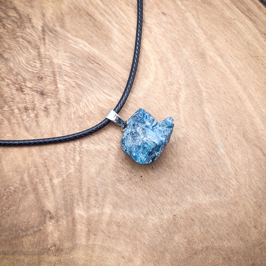 Blue Apatite raw gemstone pendant on black cord, against a wood backdrop, showcasing its vibrant blue color and natural texture.