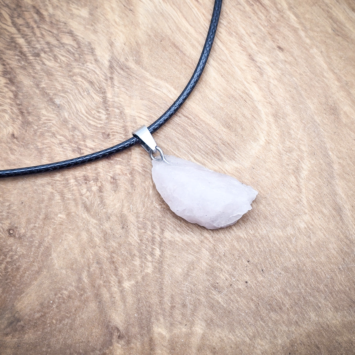 Rose Quartz raw gemstone pendant on a black cord, displayed against wood, highlighting its soft pink color and raw beauty.