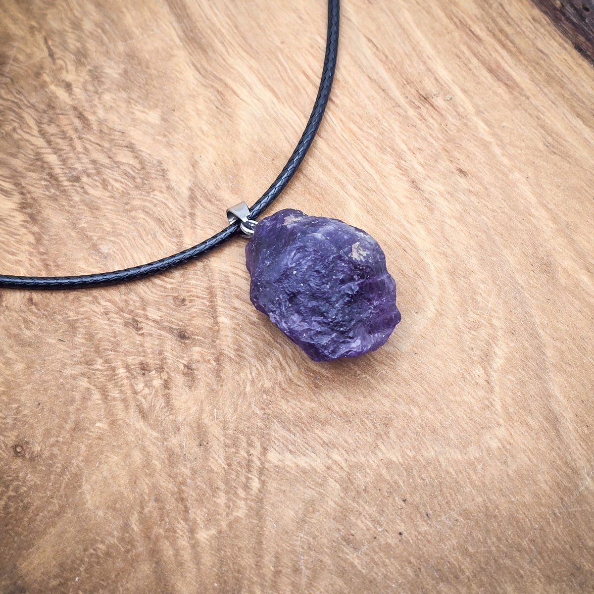 Amethyst raw gemstone pendant on a black cord, displayed on wood, showcasing its vibrant purple color and natural beauty.