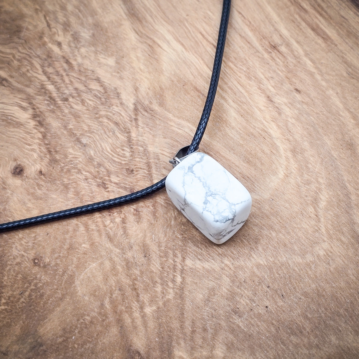 Polished Howlite cube pendant on black cord, displayed on wood, highlighting its tranquil white and grey veining.