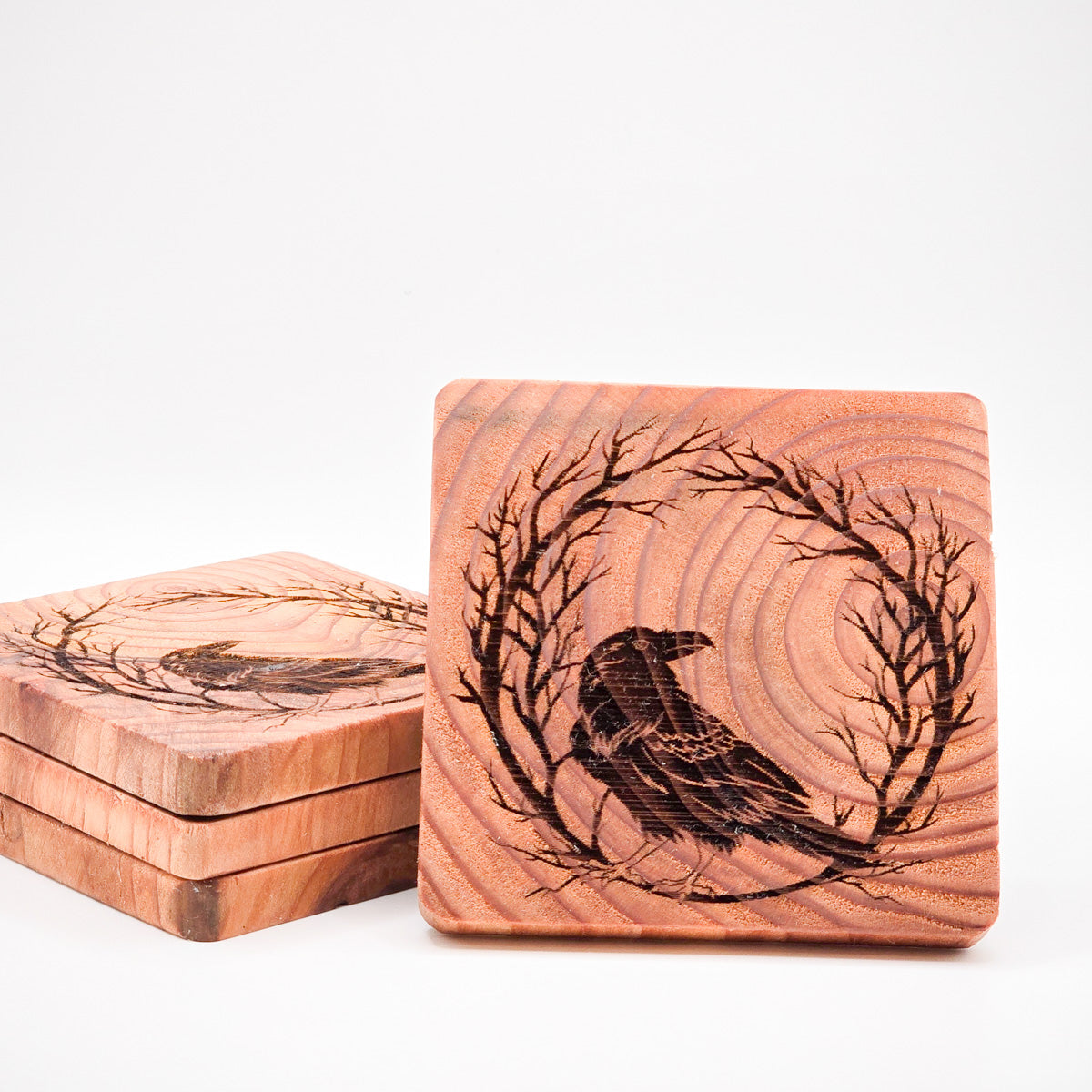 Stack of Crow on Branch Redwood Coasters with one upright, showcasing detailed artwork